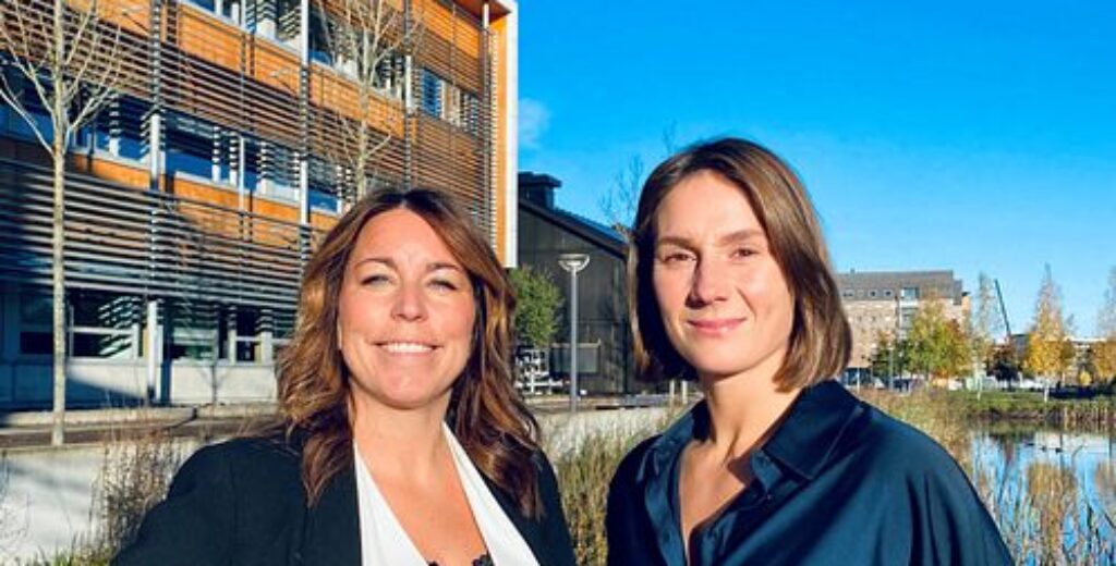 Två kvinnor från står framför en byggnad, båda ler mot kameran.