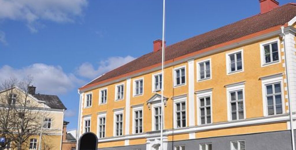 Gul byggnad med flaggstång i småstadsmiljö, blå himmel.