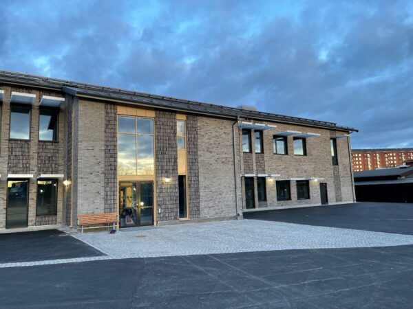 En tvåvånings skolbyggnad i ljust tegel med stora fönster och markerad glasentré står mot en dramatisk kvällshimmel; anläggningen, uppförd av GBJ Bygg, kombinerar modern materialitet, energieffektiva lösningar och en trygg gårdsyta för Brunnsboskolans verksamhet.