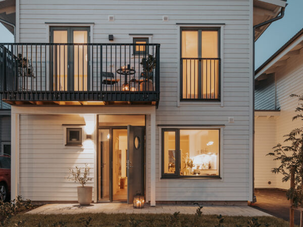 Ett tvåvåningshus i ljus träfasad lyser varmt i kvällsljuset, med balkong och stora fönster som öppnar upp mot gårdssidan; byggnaden, uppförd av GBJ Bygg, framhäver modern bostadsarkitektur med fokus på hemtrevnad, hållbara material och ett välkomnande entréparti.