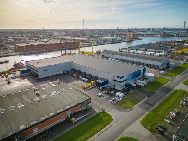 En flygbild visar ett stort logistikområde med lastbilar, lagerbyggnader och hamninfrastruktur, där GBJ Bygg har utvecklat funktionella och högkapacitativa lokaler för Mileway Söderarm, strategiskt placerade nära transportleder och vattnet för effektiv godsflödeshantering.