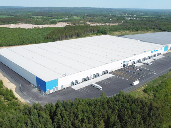 En stor logistik- och lageranläggning i vitt och blått ligger intill skog och lastzoner; byggnaden, utvecklad av GBJ Bygg, är utformad för Biltemas högvolymslogistik med effektiv åtkomst, omfattande angöringsytor och tydlig struktur.