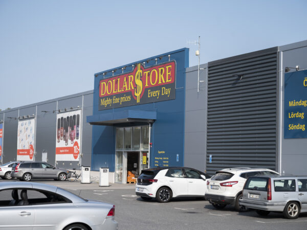 En stor handelsbyggnad med Dollarstores färgstarka entréskylt och bred parkeringsyta framför visar ett livligt kundflöde; anläggningen, utvecklad av GBJ Bygg, är utformad för effektiv logistik, tydlig exponering och ett välkomnande kommersiellt uttryck.