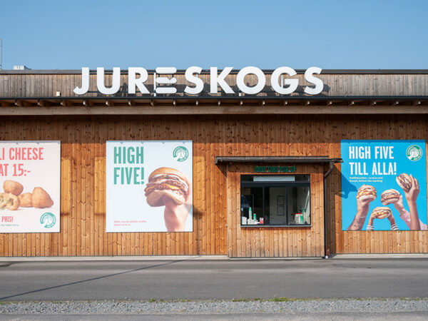 En fasad i varm träpanel med stora kampanjaffischer och tydlig Jureskogs-skylt visas mot gatan; byggnaden, anpassad och färdigställd av GBJ Bygg, kombinerar modern restaurangdesign med naturliga material för ett välkomnande och uttrycksfullt gatuläge.