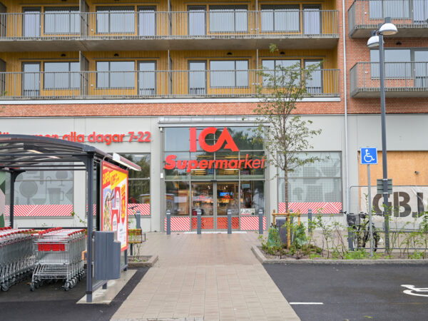 Entrén till ICA och övriga lokaler i Dalbo Centrum syns längs ett gångstråk med glasfasader och butiksfront; centrumdelen, utvecklad av GBJ Bygg, skapar ett välkomnande och lättillgängligt nav för stadsdelens service.