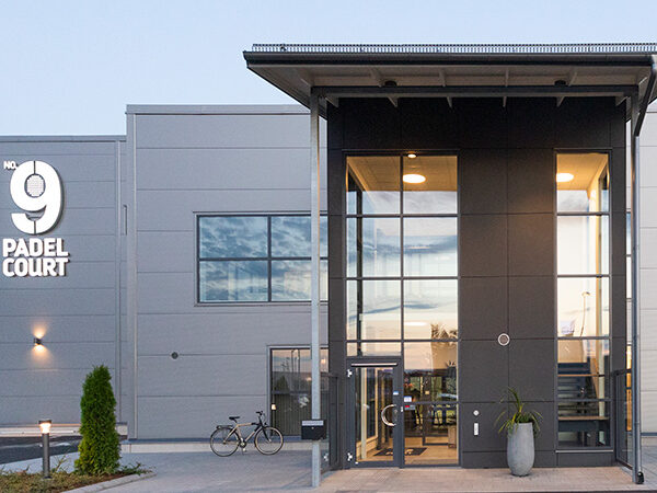 En modern padelanläggning med glasad entré och stora fasadpartier står upplyst i kvällsljuset; byggnaden, framtagen av GBJ Bygg, kombinerar robust industrikänsla med ett välkomnande uttryck där skylten No. 9 Padel Court markerar anläggningens identitet.