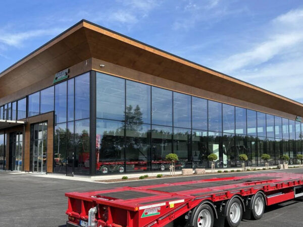 En modern industribyggnad med stora glasfasader och karaktäristiskt utskjutande tak syns i solljus; anläggningen, uppförd av GBJ Bygg, kombinerar transparent arkitektur, robust konstruktion och flexibla ytor anpassade för Hakarps Equipment och deras maskinrelaterade verksamhet, med ett rött trailerflak i förgrunden.