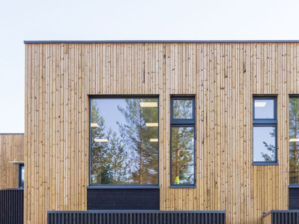 En modern skolbyggnad i trä med stora fönsterpartier står mot en ljus himmel; fasaden, uppförd av GBJ Bygg, kombinerar hållbart materialval med tydlig struktur och skapar en varm och inbjudande miljö för Flesbergskolans elever och personal.