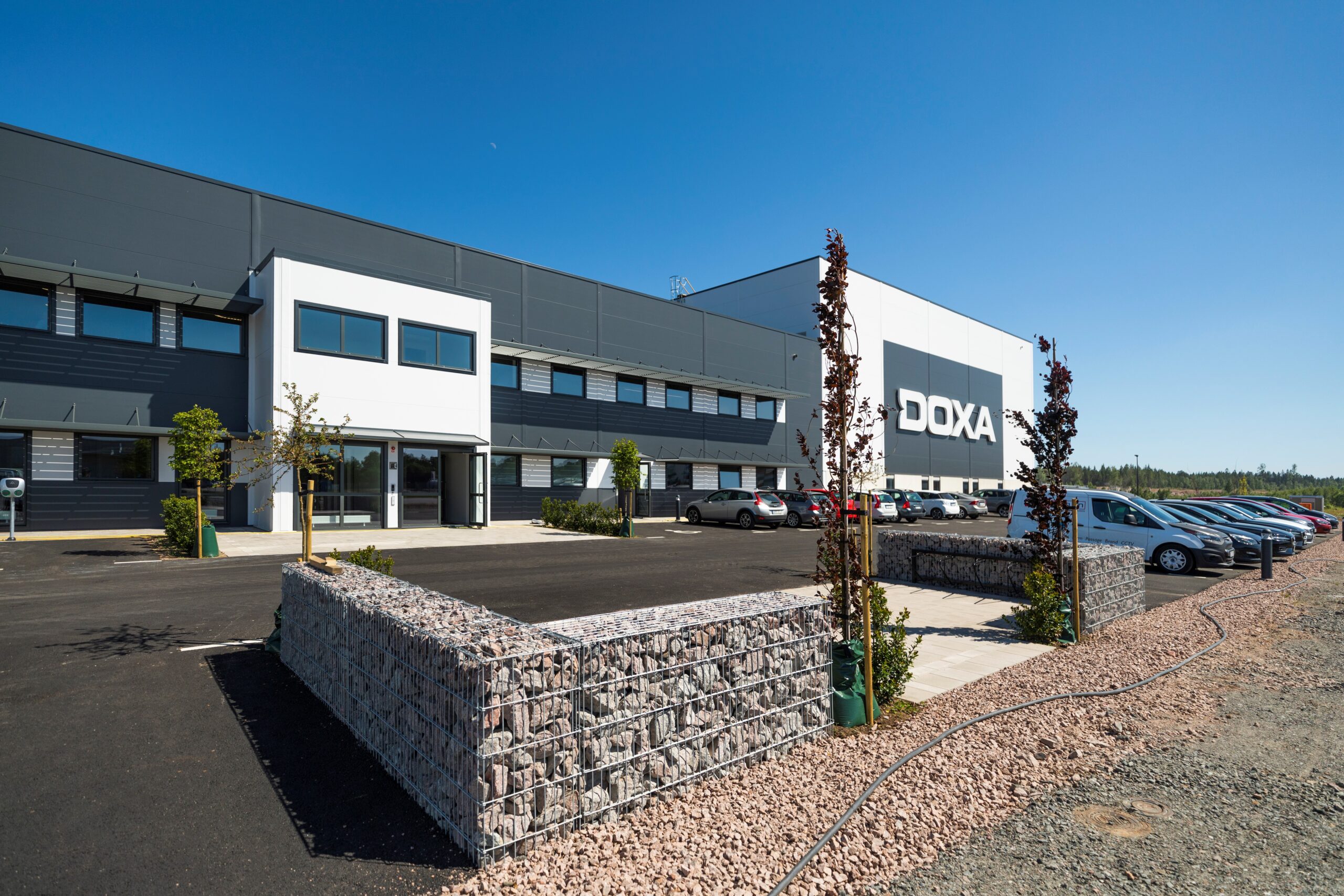 En modern industribyggnad med vit och mörkgrå fasad står längs en nyanlagd parkeringsyta, där Doxa-logotypen framträder tydligt; anläggningen, utvecklad av GBJ Bygg, kombinerar funktionella logistiklösningar med en representativ och stilren företagsentré.