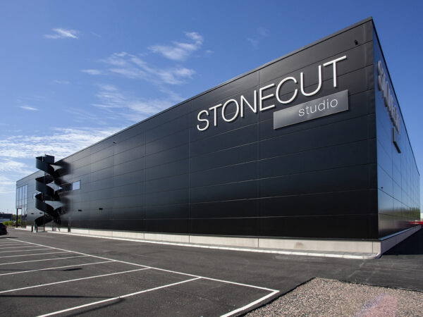 En stor och modern industribyggnad med svart fasad och tydlig skyltning STONECUT studio står mot en klar blå himmel; byggnaden, uppförd av GBJ Bygg, har rena linjer, markerad brandtrappa och rymliga parkeringsytor som understryker dess funktionella och stilrena utformning.