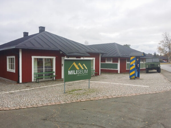 Ett traditionellt rött hus med svart sadeltak står intill en stenlagd gårdsplan där skylten Miliseum Skillingaryd är placerad; byggnaden, renoverad och anpassad av GBJ Bygg, kombinerar kulturhistoriska uttryck med funktionella ytor för museets verksamhet, kompletterat av militärfordon och vaktsymbolik i miljön.