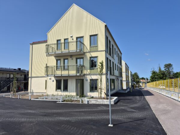 Ett modernt bostadshus i tre våningar med sadeltak och vertikal träpanel, byggt av GBJ Bygg som del av bostadsprojektet Färjebacken, där balkonger, energieffektiva fönster och hållbara material präglar helhetsintrycket.
