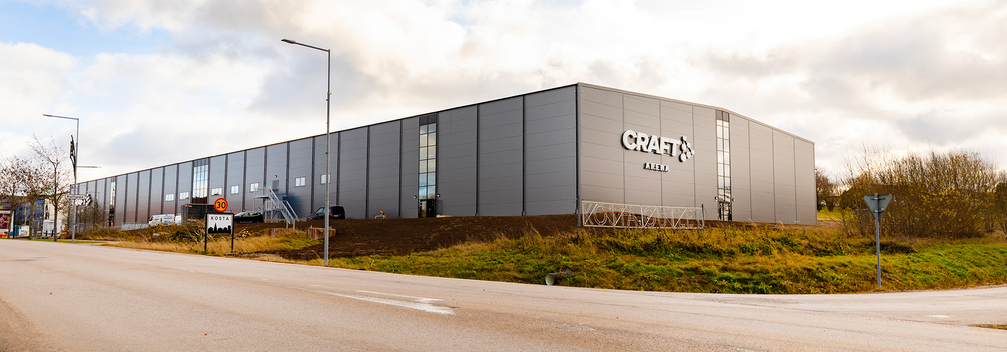 En stor modern arenabyggnad med grå fasad och tydlig skyltning för Craft Arena sträcker sig längs en väg, där GBJ Bygg har uppfört anläggningen som en funktionell och robust idrotts- och evenemangsmiljö i Kosta med fokus på hållbar konstruktion.