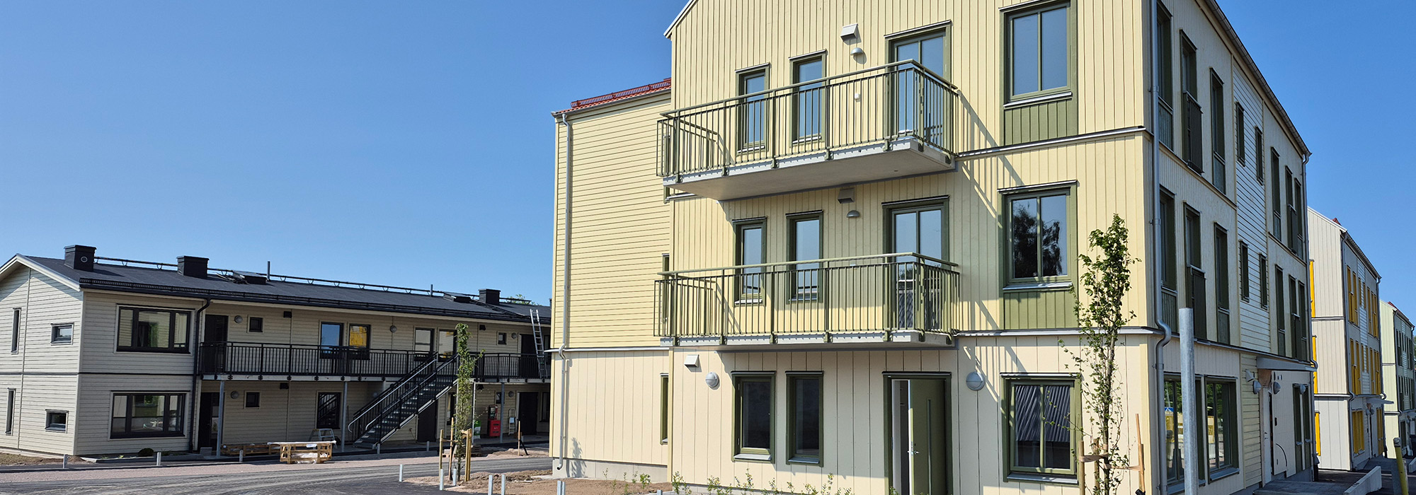 Ett nyproducerat flerbostadshus vid Färjebacken, där GBJ Bygg har uppfört bostäder i trä med ljusgul fasad, generösa balkonger och tydlig gaturumsanknytning, omgiven av nyanlagda gångytor och planteringar.