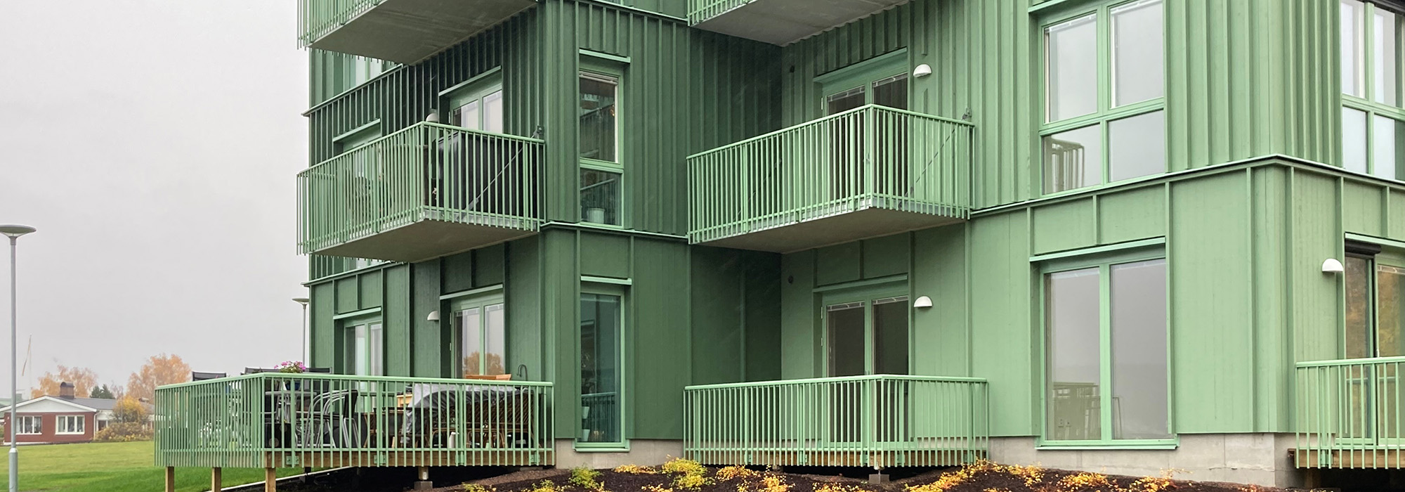 En modern flerbostadsbyggnad vid Sannaängen fasad med ljusgröna träfasader och utskjutande balkonger, där GBJ Bygg står bakom projektets hållbara utformning. Byggnaden ramas in av en nylagd gårdsmiljö och stora fönster som förstärker det öppna uttrycket.
