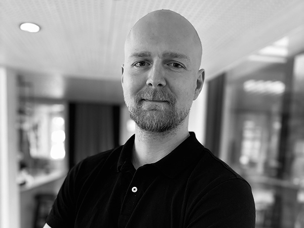 Ett porträtt i personal-linus-eriksson.jpg visar en medarbetare från GBJ Bygg som står med armarna i kors inomhus, fotograferad i kontorsmiljö med mjukt ljus, suddig bakgrund och professionellt, lugnt uttryck