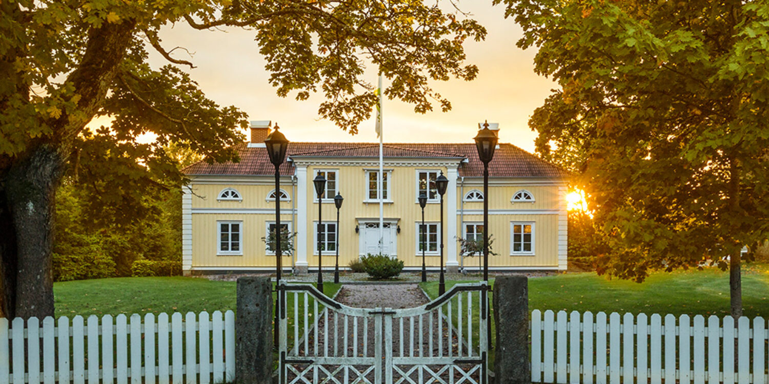 Thelestads herrgårds gula träfasad ramas in av stora lövträd, vit grind och grusgång i varmt kvällsljus. Den historiska byggnaden används som del av GBJ byggs huvudkontor och bevarar områdets traditionella herrgårdskaraktär.