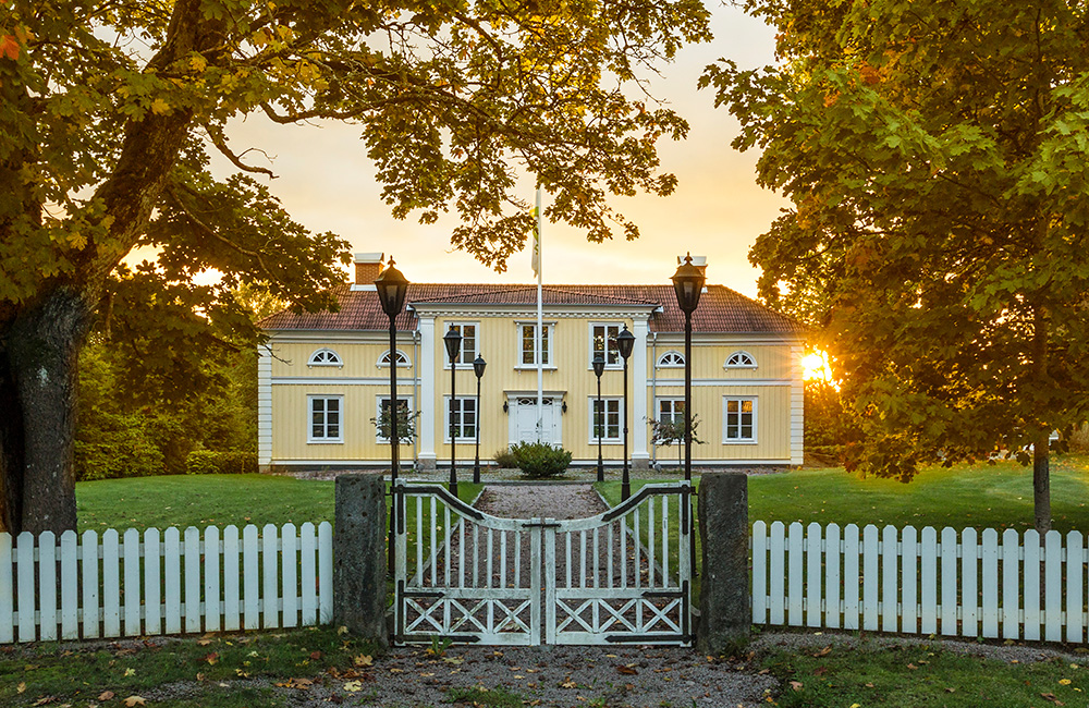 Thelestads herrgårds gula träfasad ramas in av stora lövträd, vit grind och grusgång i varmt kvällsljus. Den historiska byggnaden används som del av GBJ byggs huvudkontor och bevarar områdets traditionella herrgårdskaraktär.