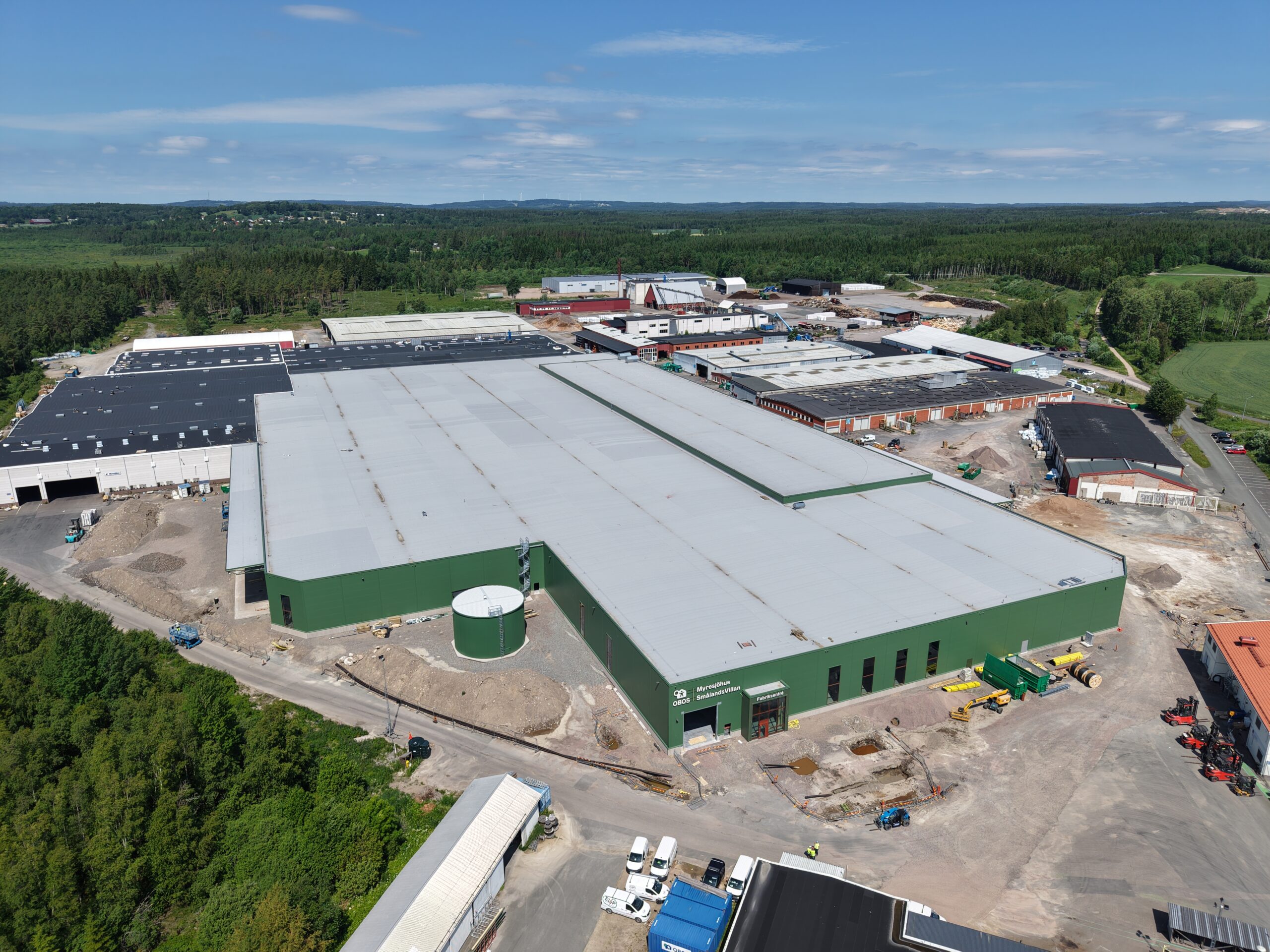 En drönarvy visar OBOS stora husfabrik med grön fasad och omfattande produktionsytor, omgiven av skog och befintliga industribyggnader. GBJ Bygg genomför utbyggnaden med pågående markarbeten runt anläggningen. Text: ”OBOS”, ”Myresjöhus”, ”SmålandsVillan”, ”Fabriksentré”