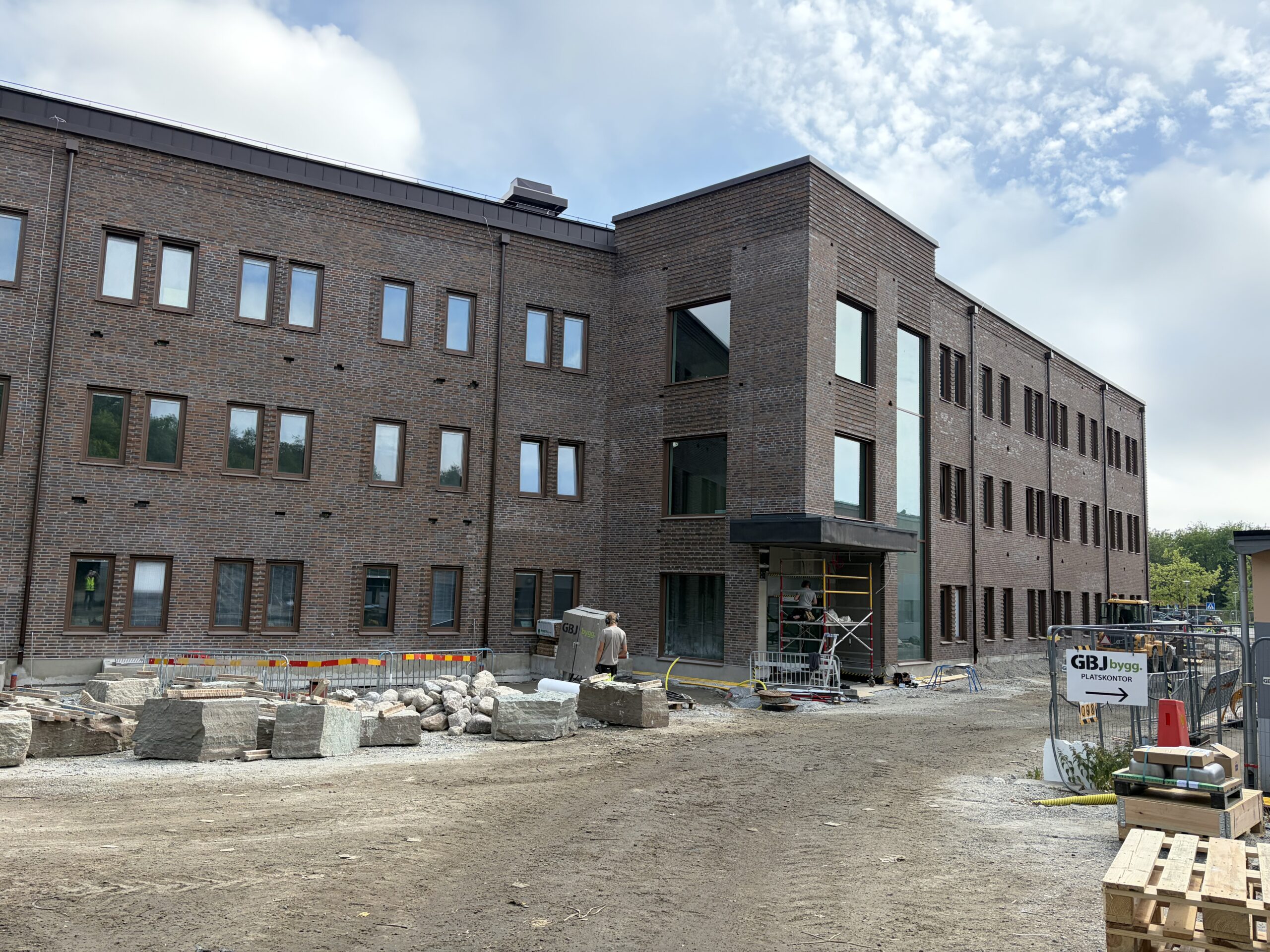 En större tegelbyggnad vid Blekingesjukhuset utvecklas med höga glaspartier kring entrén och pågående markarbeten framför fasaden. GBJ Bygg genomför projektet med fokus på att förena befintlig tegelarkitektur med moderna tillägg.