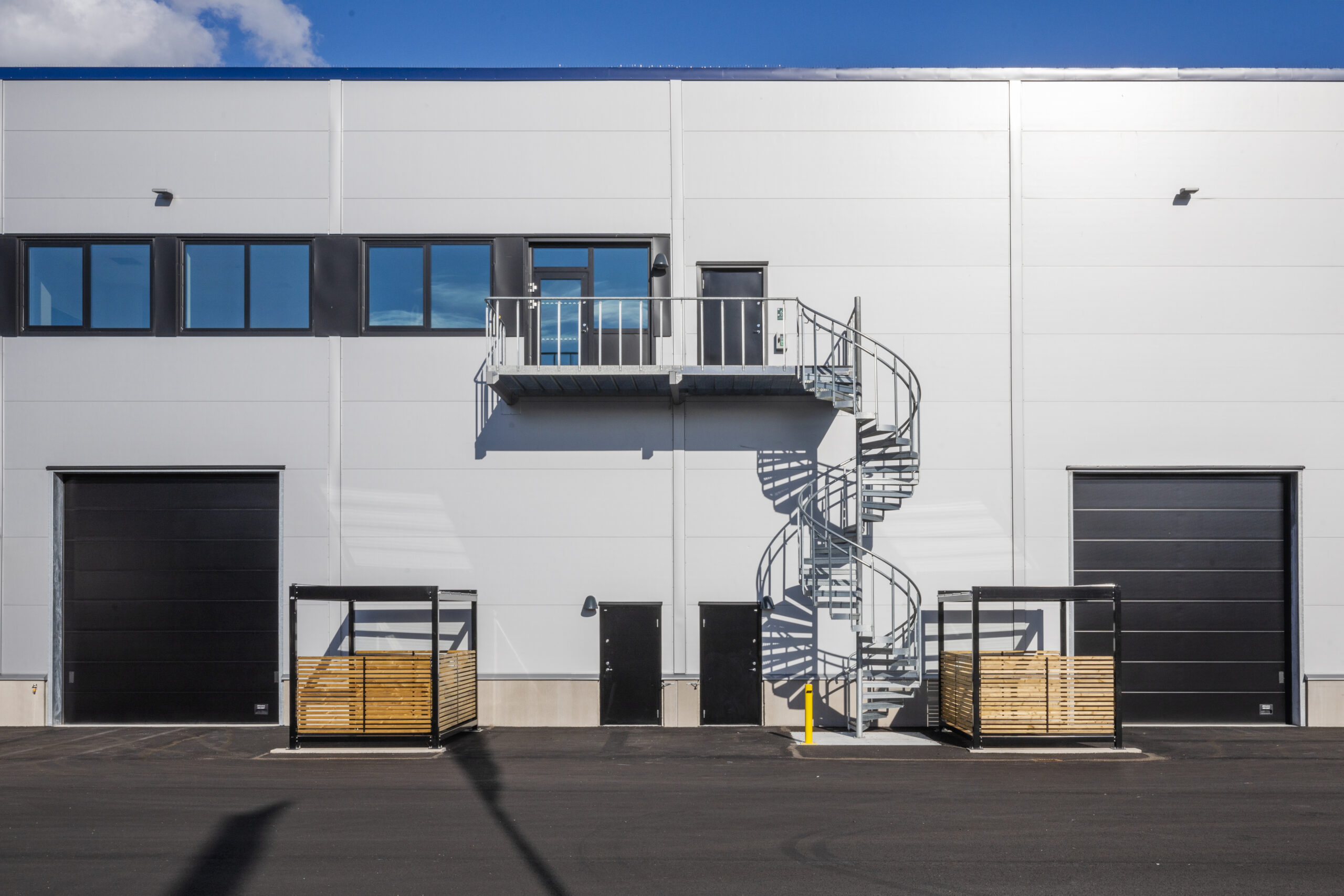 Ljus industrifasad med två svarta portar, fönster, träklädda avskärmningar och en utvändig spiraltrappa i metall. I GBJ byggs projekt i Norrköping skapar detaljerna funktionella anslutningar mellan byggnadens olika våningsplan och verksamhetsytor.