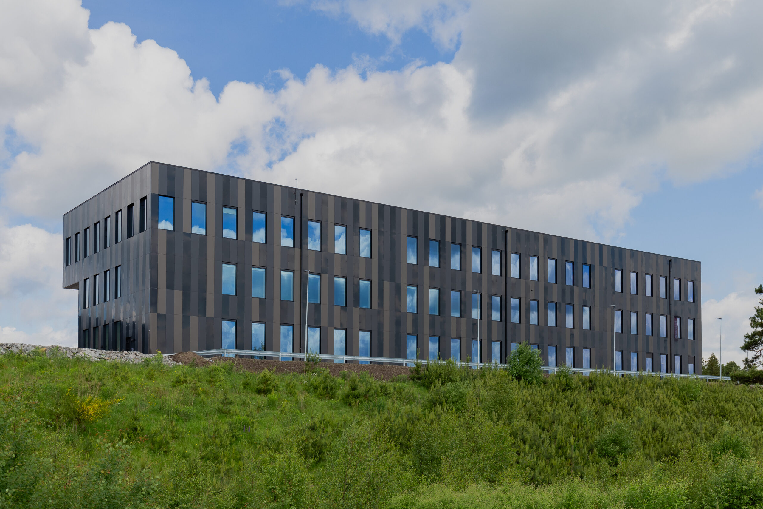 En långsträckt kontorsbyggnad i tre våningar reser sig bakom ett grönskande släntparti. GBJ Byggs projekt präglas av en mörk, varierad fasadbeklädnad och regelbundet placerade fönster som ger byggnaden ett modernt uttryck.