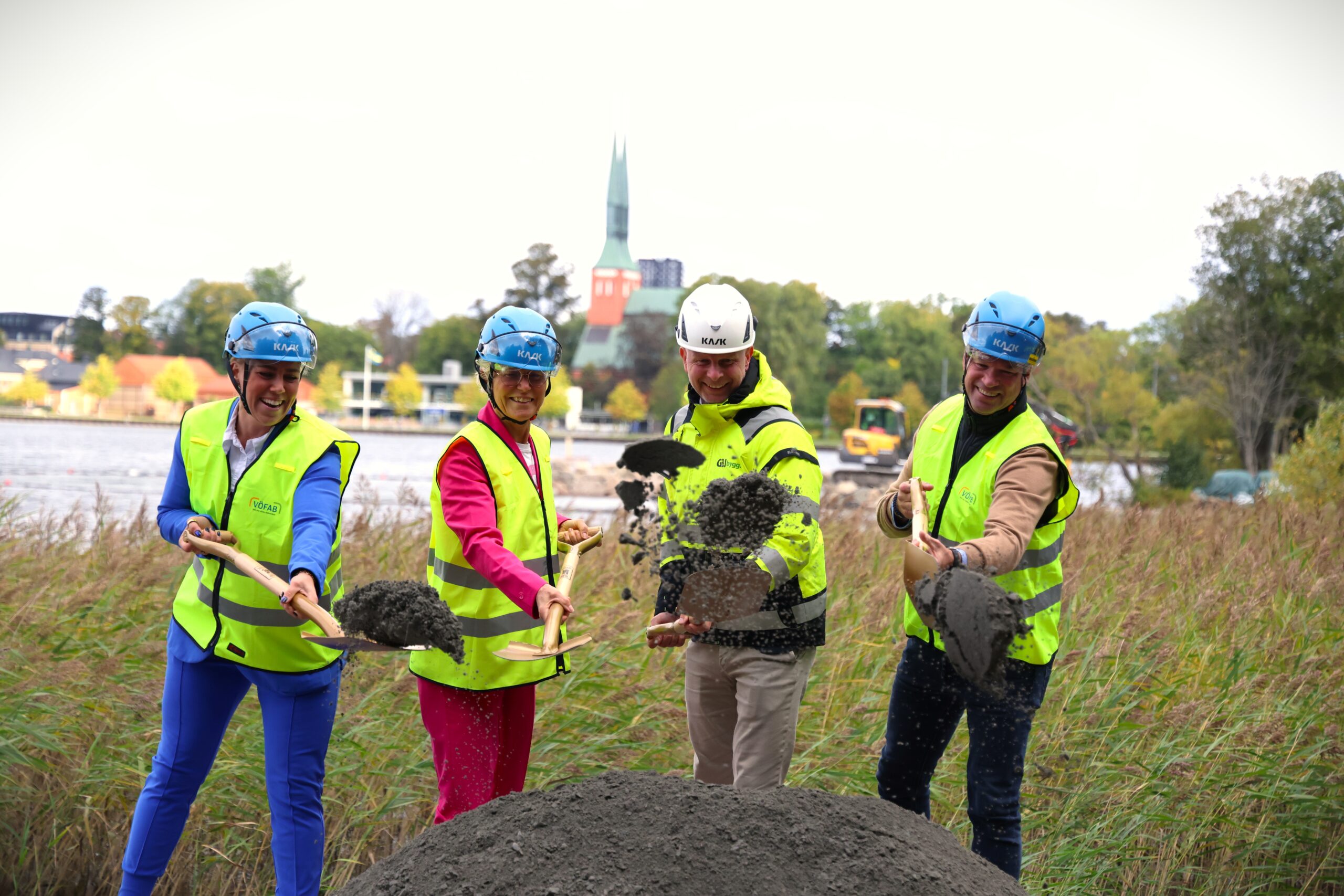 Fyra personer i hjälmar och reflexvästar tar det första spadtaget för ett nytt kallbadhus i Växjö. Med Växjösjön och domkyrkan i bakgrunden markerar ceremonin starten på GBJ Byggs byggprojekt.