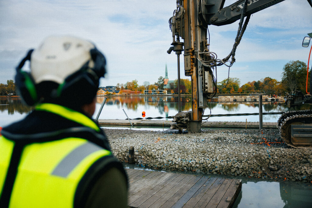En platschef från GBJ bygg övervakar pålningsarbetet för det nya kallbadhuset vid Växjösjön. I förgrunden syns hans skyddshjälm och reflexväst, medan en stor pålningsmaskin arbetar på den stenfyllda strandkanten med Växjö domkyrka i bakgrunden.