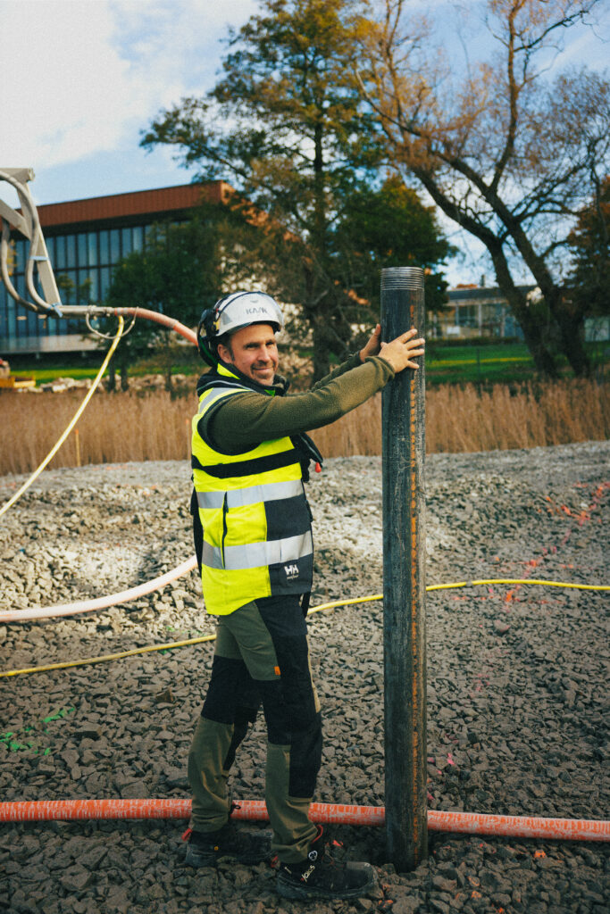En medarbetare från GBJ bygg håller i en stålpåle under grundläggningsarbetet för kallbadhuset i Växjö. Han bär skyddshjälm och reflexväst och står på en grusbelagd arbetsyta med byggnader och träd i bakgrunden.