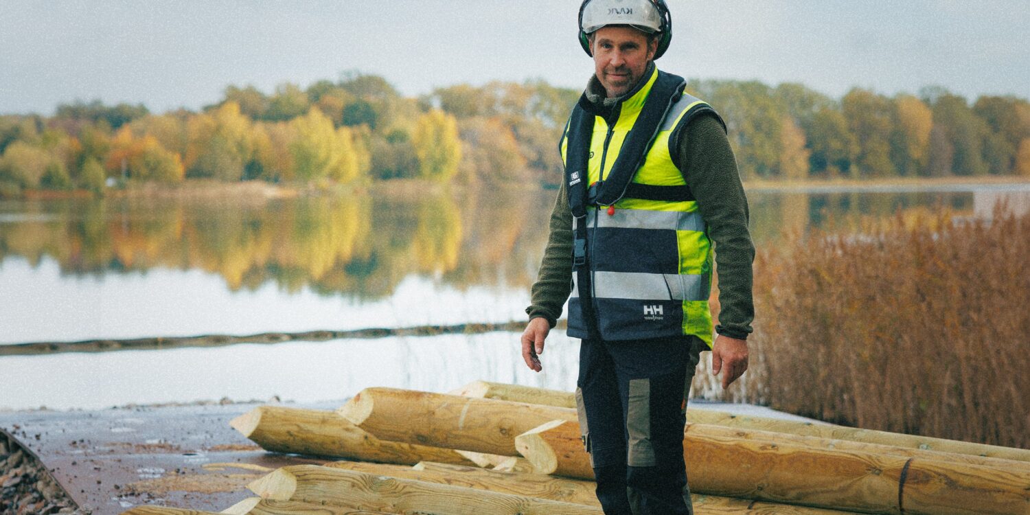 En byggarbetare från GBJ bygg står vid träpålar som lagts ut på en stenbelagd strand inför arbetet med kallbadhuset. Han bär hjälm, hörselskydd och varselväst, med en stilla sjö och höstfärgade träd i bakgrunden.